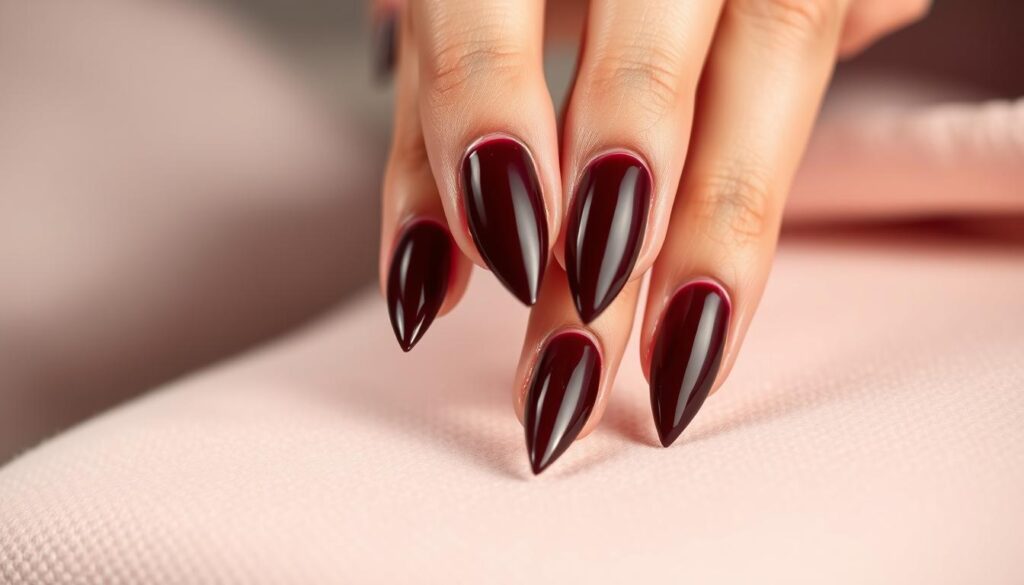 A beautifully lit close-up of neatly manicured stiletto nails, set against a soft, blurred background. The nails are perfectly filed to a sharp, dramatic point, their glossy, deep burgundy polish reflecting the warm lighting. The cuticles are neatly trimmed, and a high-shine top coat adds a luxurious, glass-like finish. Elegant yet practical, the image showcases the careful attention to detail required for impeccable stiletto nail care.